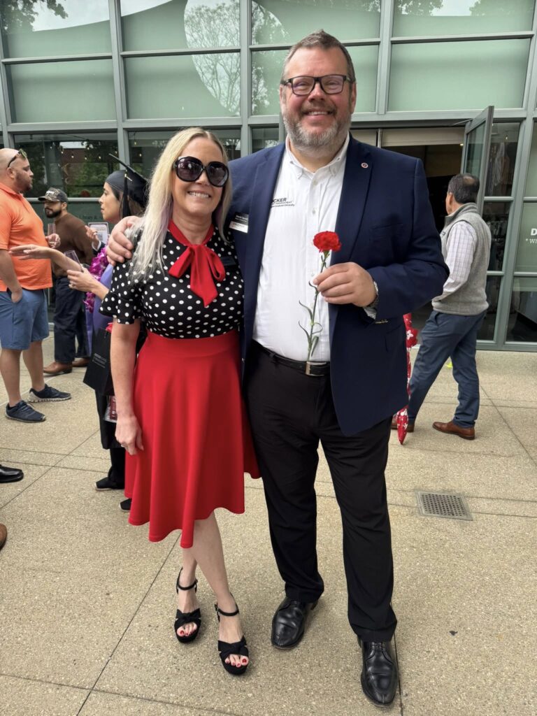a woman with blonde hair stands next to a man in a suit. he is holding a rose and she is wearing sunglasses near a CGU building